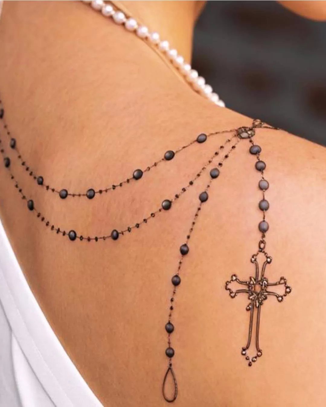 Ornate black ink cross tattoo with beaded chains draped across a woman's shoulder.