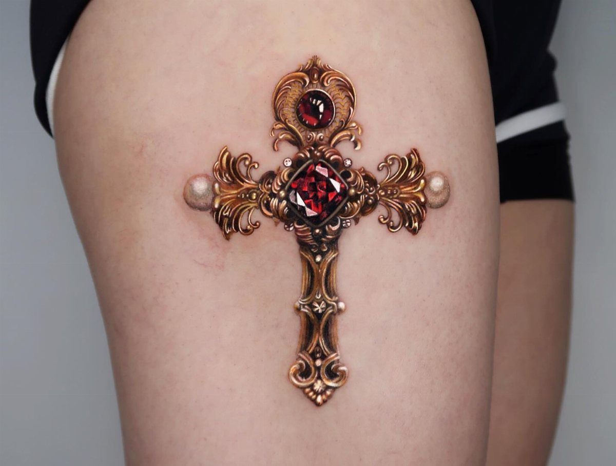 Ornate bronze cross tattoo with red gemstones and pearls on a person's thigh