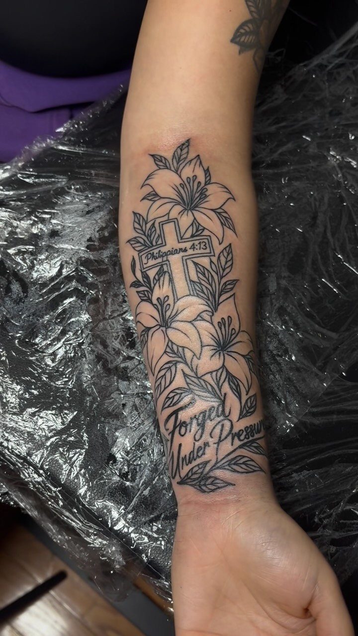 Black ink tattoo of a cross surrounded by lilies on a forearm