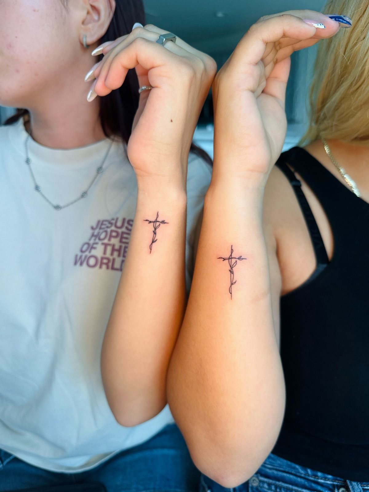 Two inner wrists with small black ink cross tattoos entwined with delicate vines.