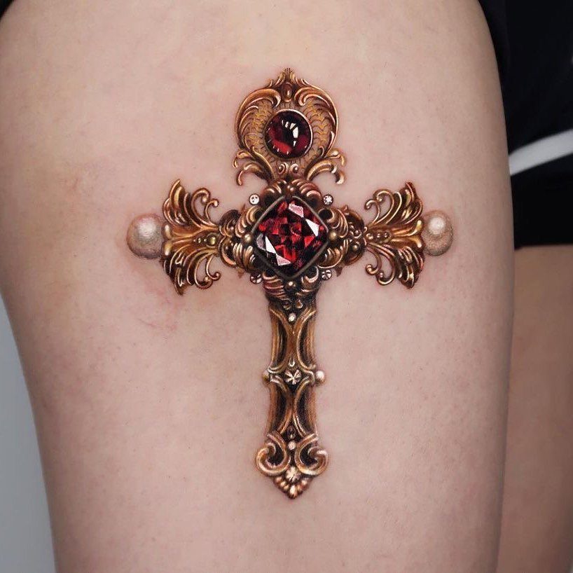 Ornate metallic gold cross tattoo with red jewels on a person's skin.