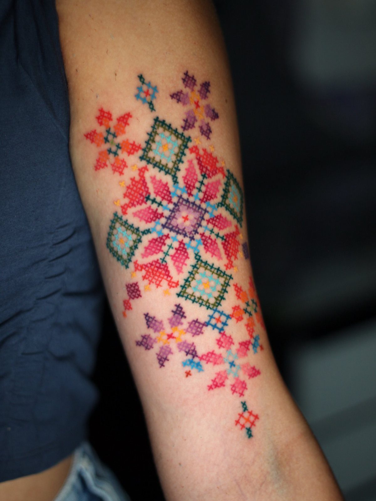 Ornate colorful cross tattoo with geometric cross-stitch patterns on a person's upper arm.
