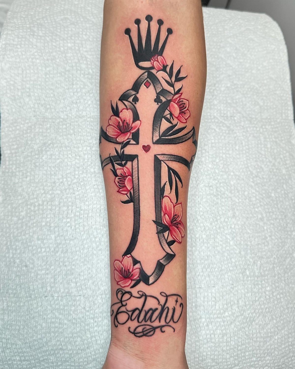 Ornate black cross tattoo with pink flowers and a crown on a forearm