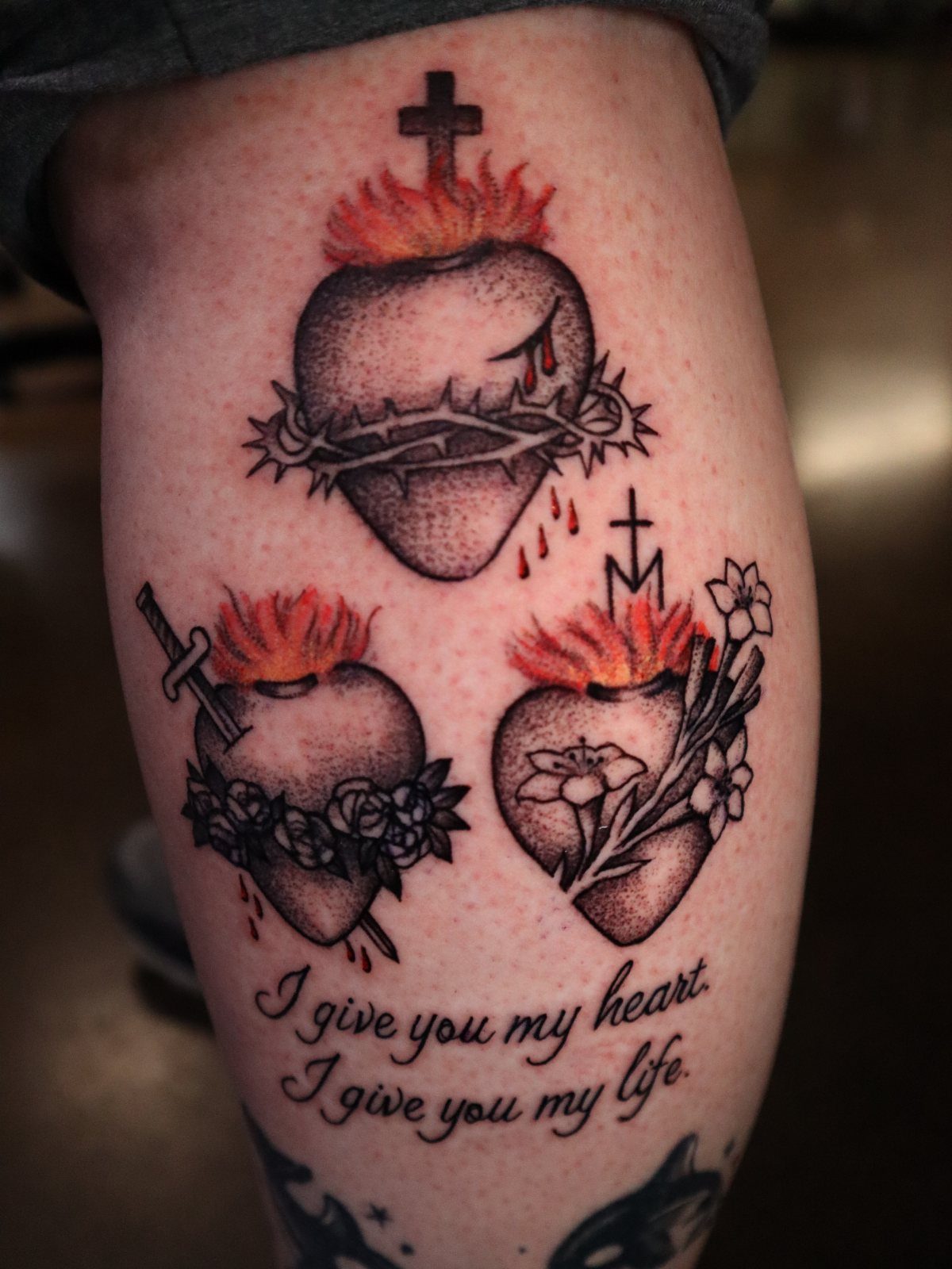 Three sacred heart tattoos with crosses, flames, and flowers on a person's leg.