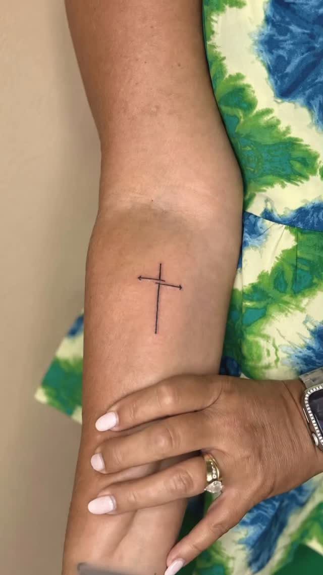 Small simple black cross tattoo on a person's forearm with manicured hand.