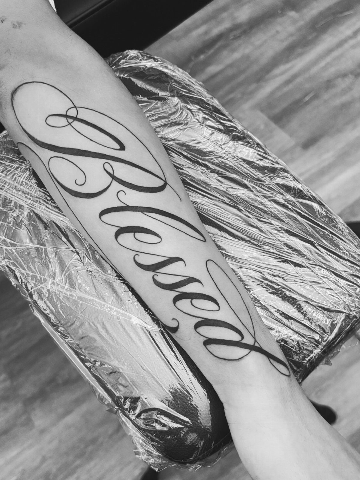 Cursive tattoo reading Blessed on a forearm resting on plastic wrap.