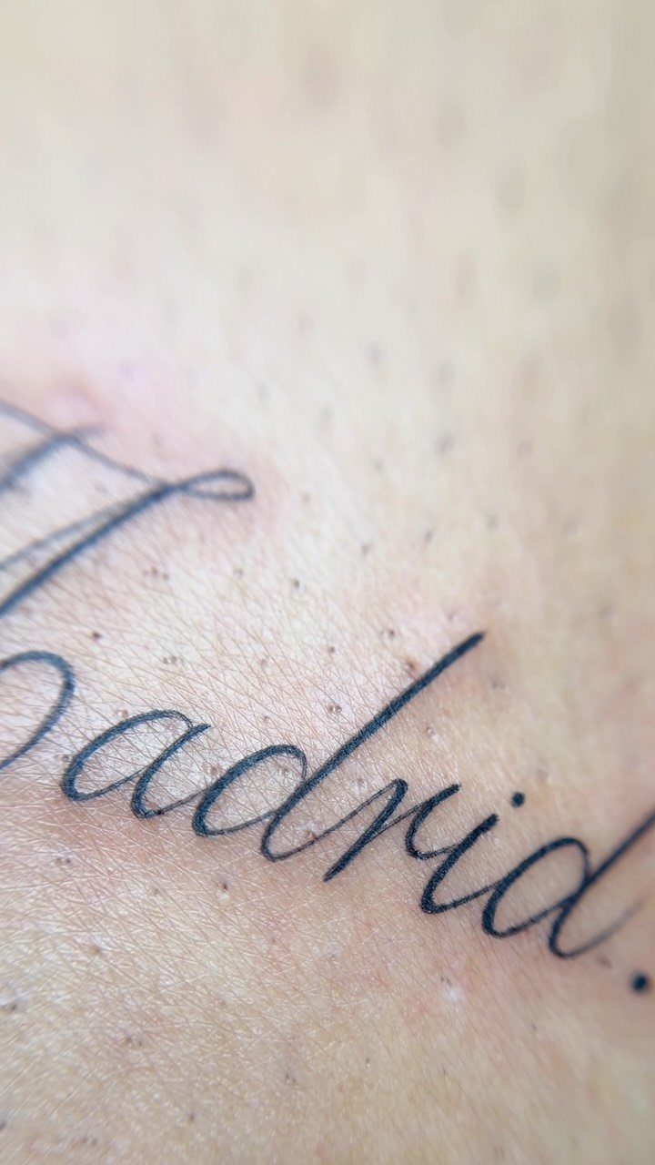 Cursive tattoo of the word Madrid in black ink on fair skin.