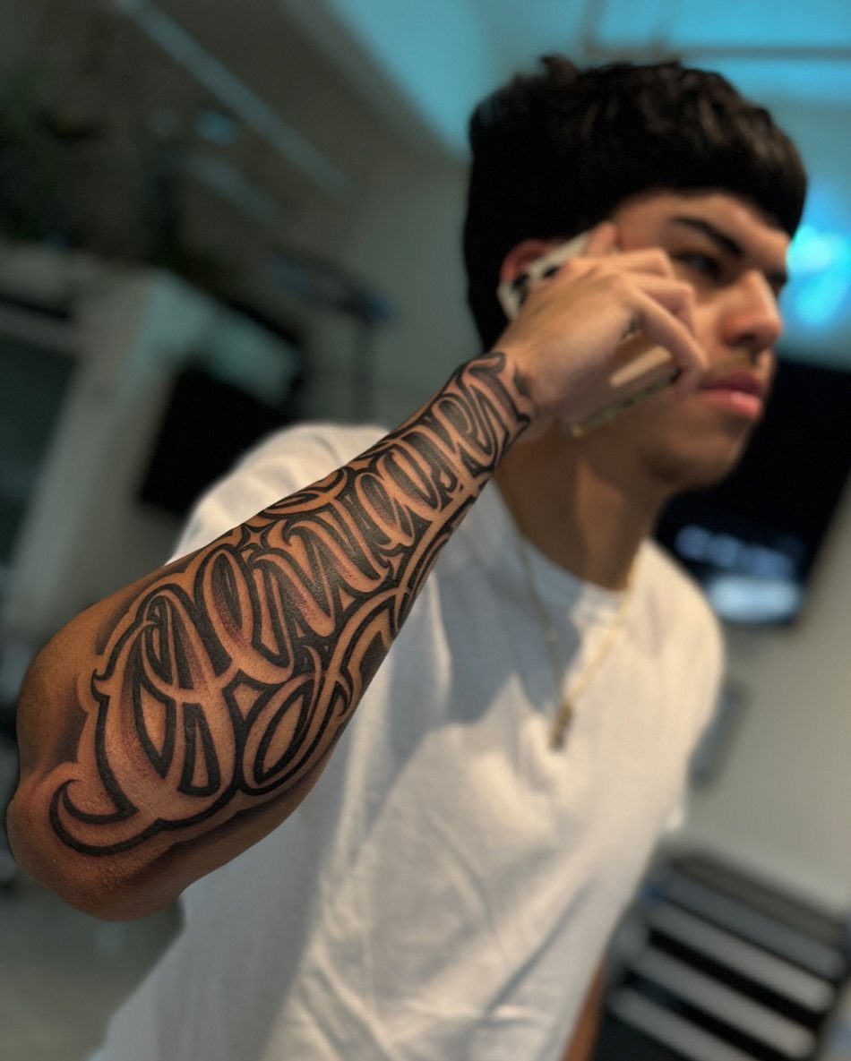 Man with a large black cursive lettering tattoo on his forearm and wrist.