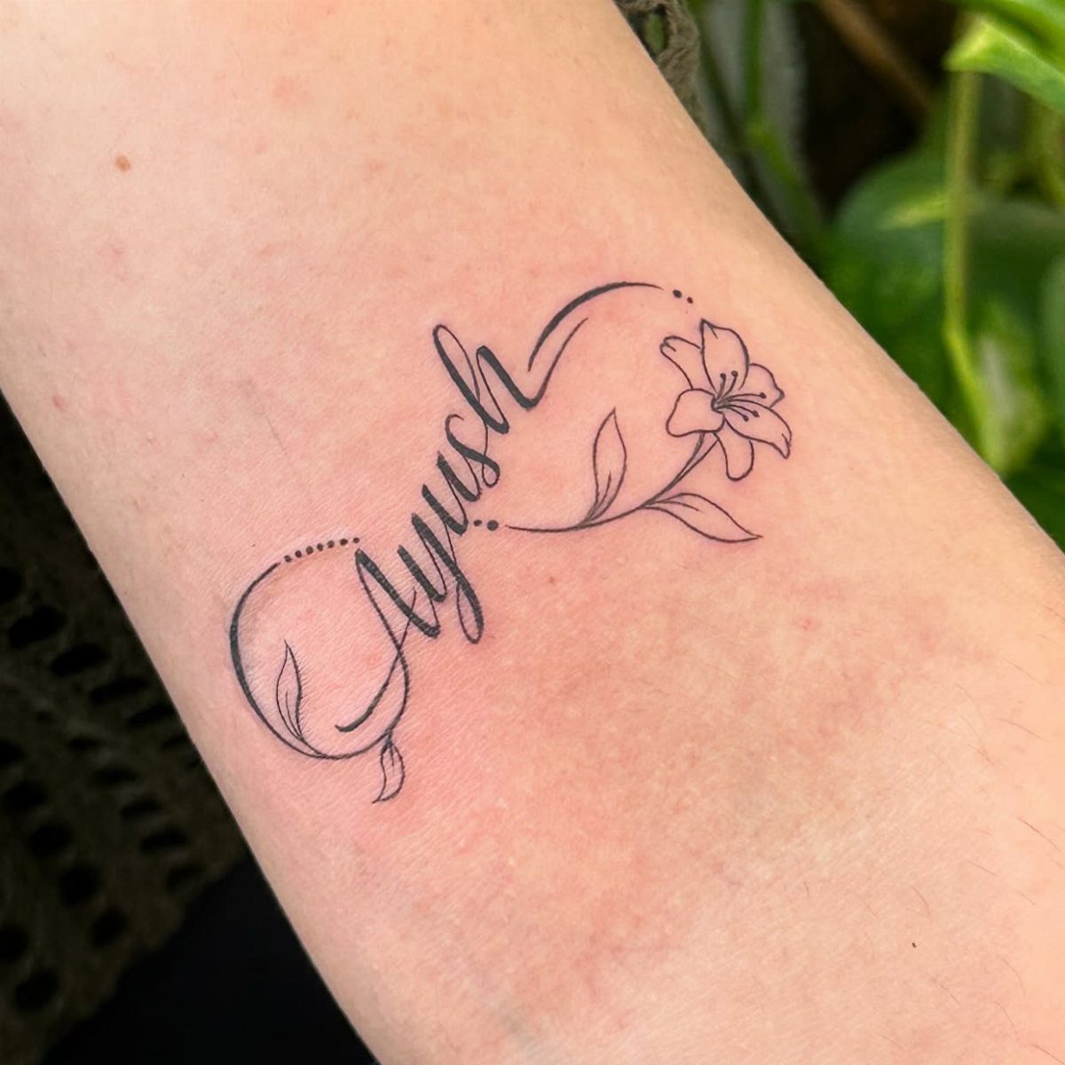 Cursive name tattoo of Ayush with a small flower on a forearm