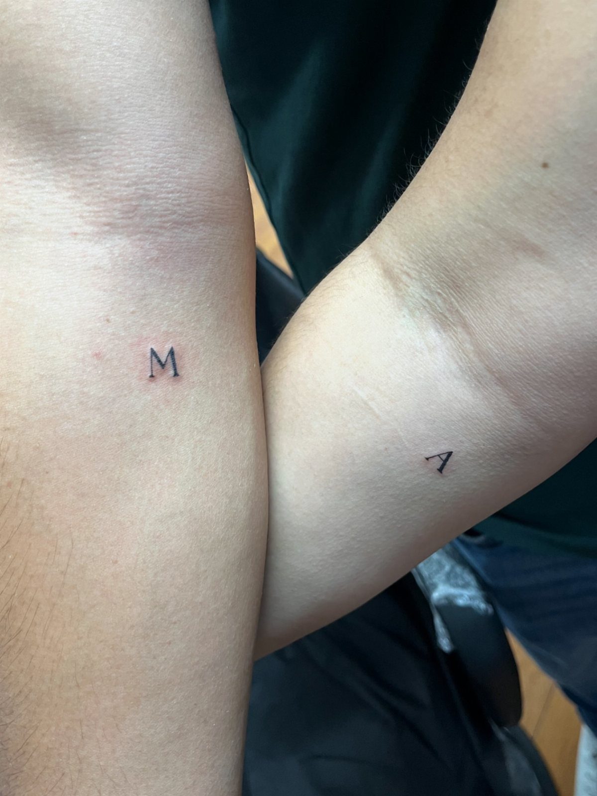 Two inner forearms with small black ink initial tattoos of M and A.