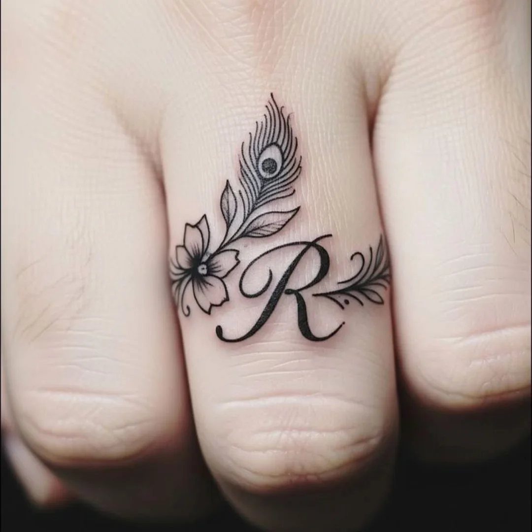 Cursive letter R tattoo on a finger with a flower and peacock feather.