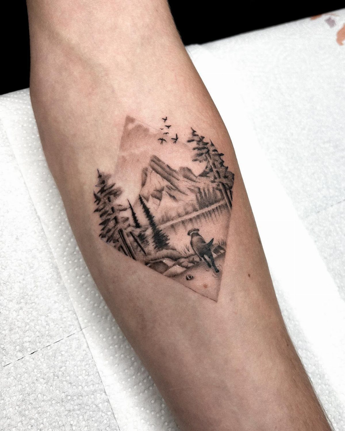 Landscape tattoo on forearm featuring mountains, pine trees, and a small dog.