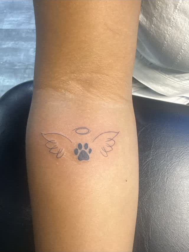 Dog paw print tattoo with wings and a halo on a forearm.