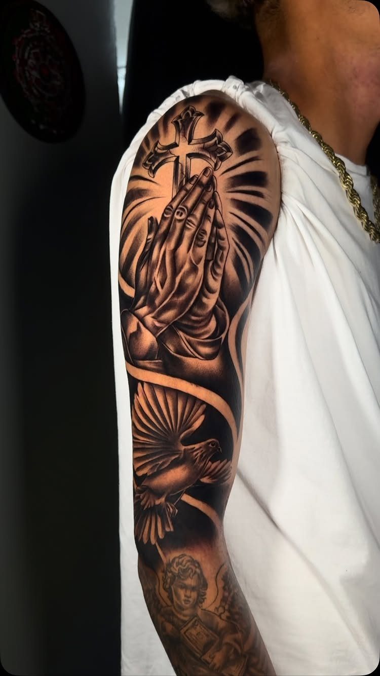 Praying hands and a cross tattoo on a man's upper arm with a dove.