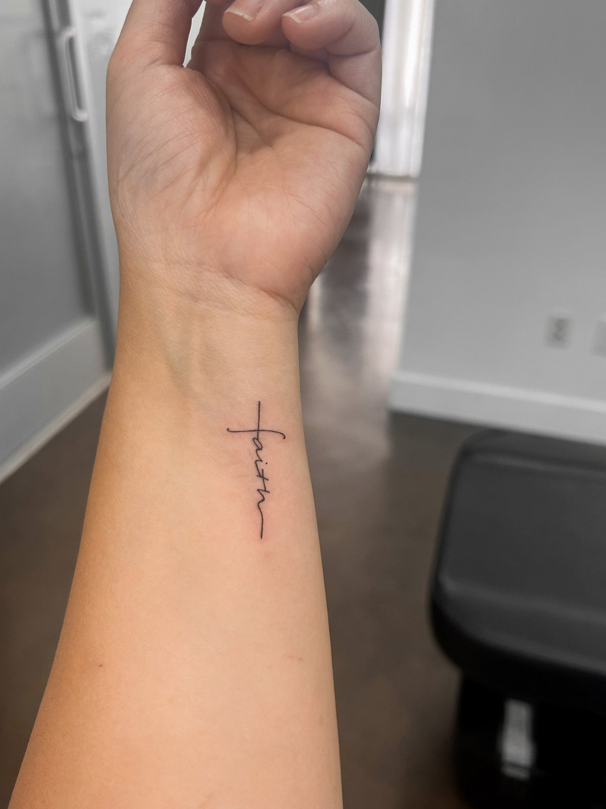 Small black ink tattoo of a cross with faith written in cursive on wrist