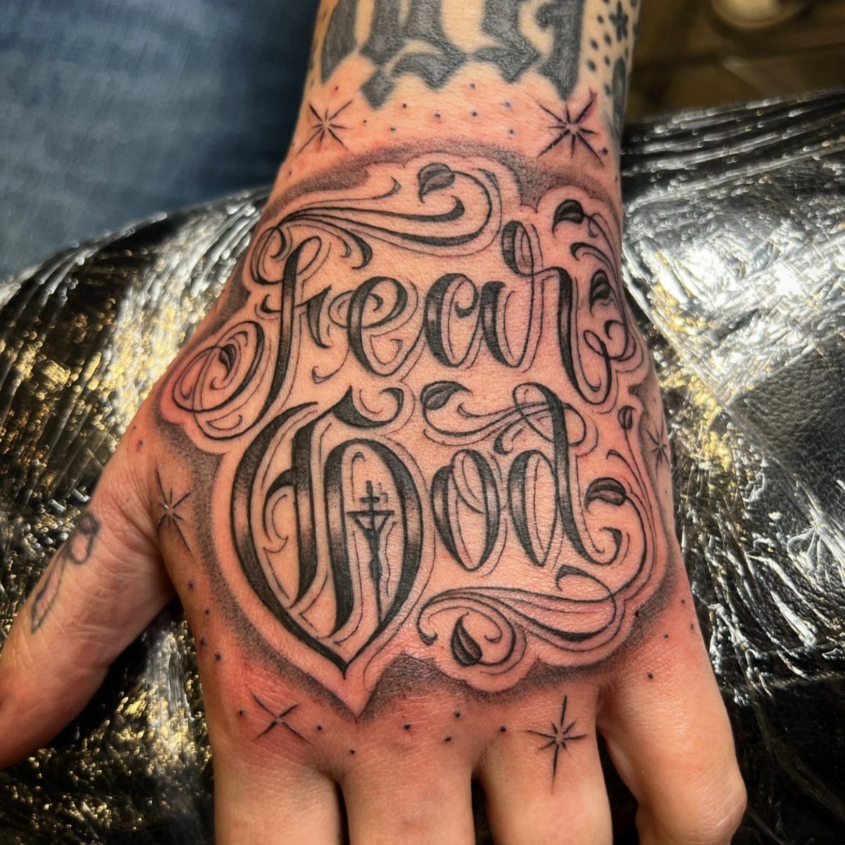 Ornate black ink tattoo on a hand reading Fear God in calligraphic script.