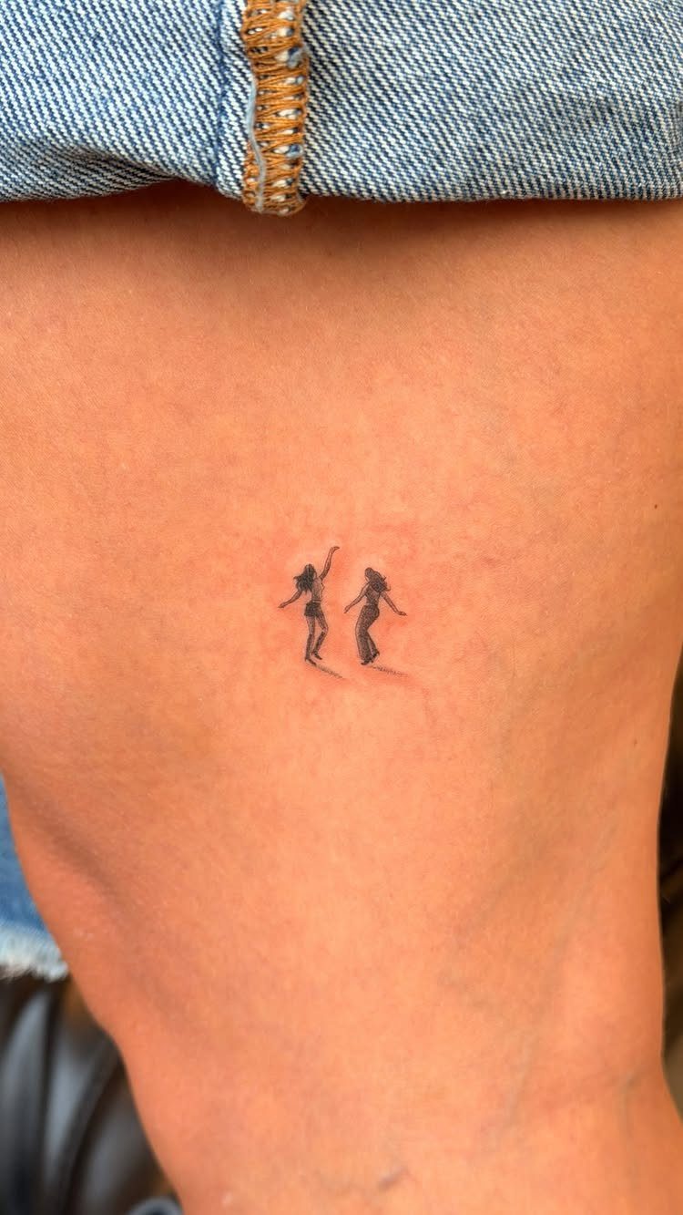 Small black ink tattoo of two dancing female silhouettes on a person's leg.