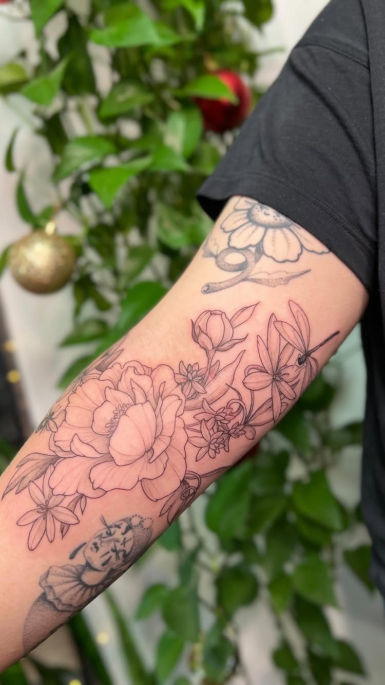 Floral fine line tattoos on a person's arm against a background of green leaves.