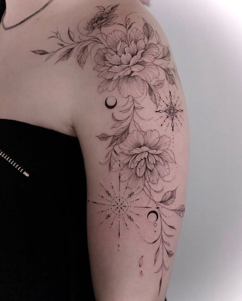 Floral tattoo with peonies and stars in thin black lines on an upper arm.