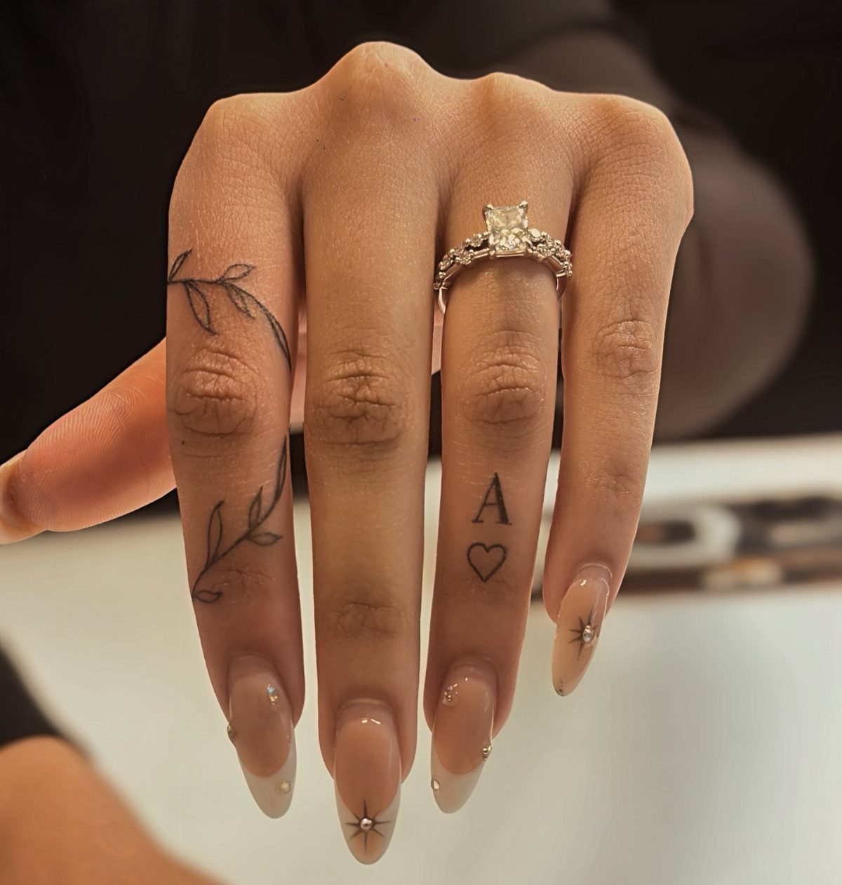 Hand with manicured nails, a diamond ring, and finger tattoos of a vine and initial.