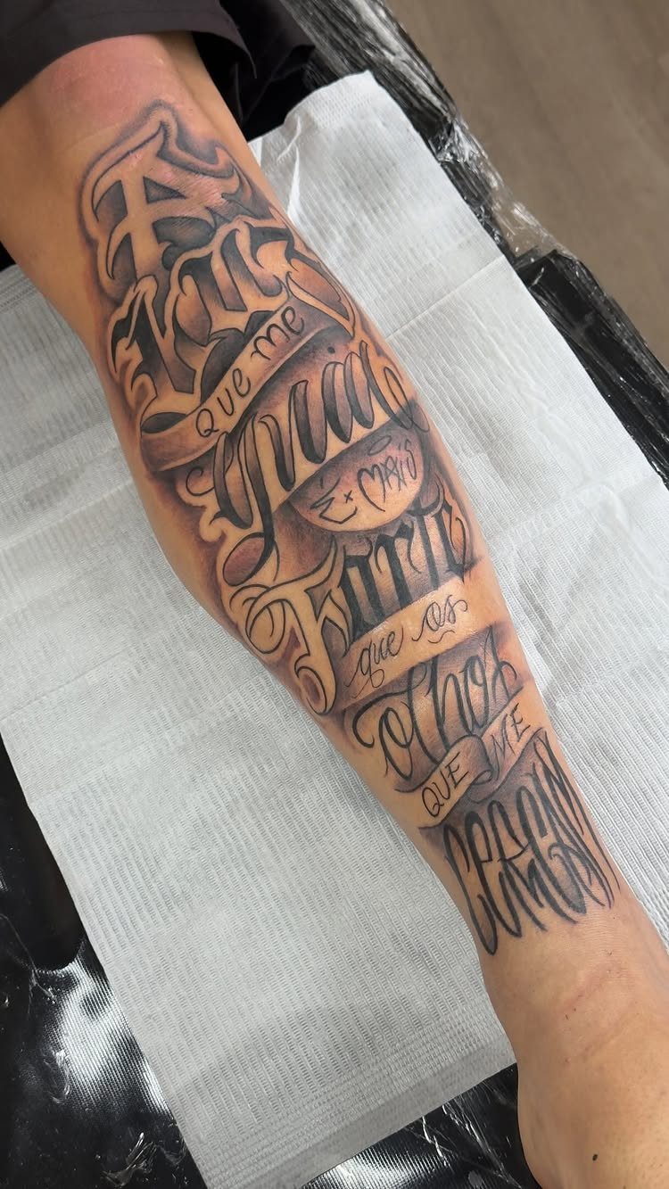 Ornate Chicano style script tattoo on a person's forearm with flowing grey ink lettering.