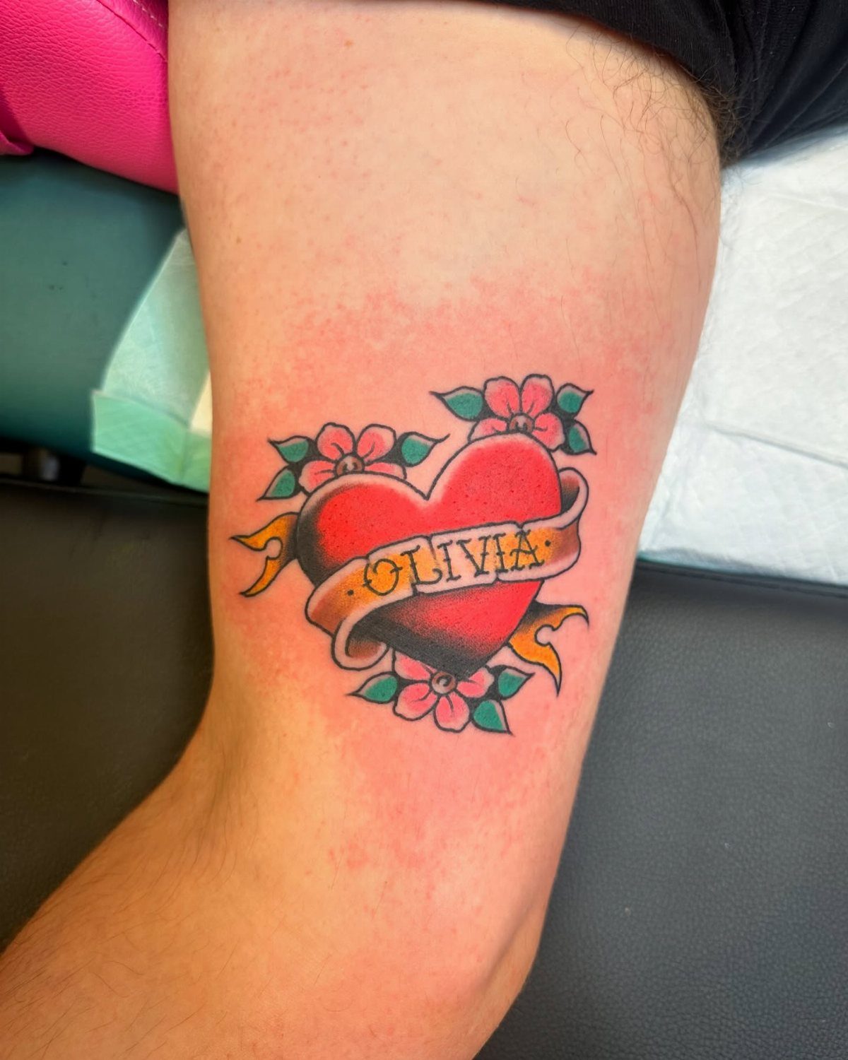 Red heart tattoo with the name Olivia on a yellow banner and pink flowers.