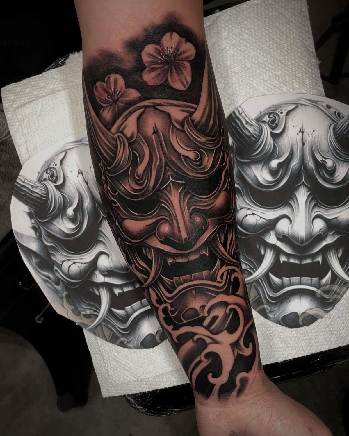 Hannya mask tattoo on a forearm with cherry blossoms and reference sketches nearby