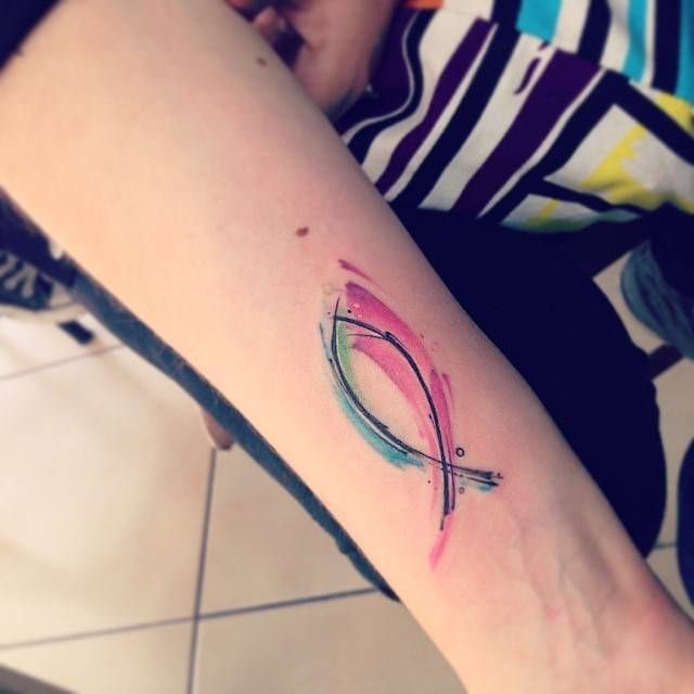 Jesus fish tattoo on a forearm with colorful pink and teal watercolor effects.