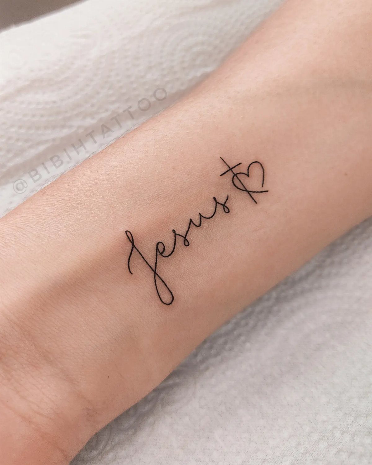 Cursive Jesus tattoo with a cross and heart on a person's forearm