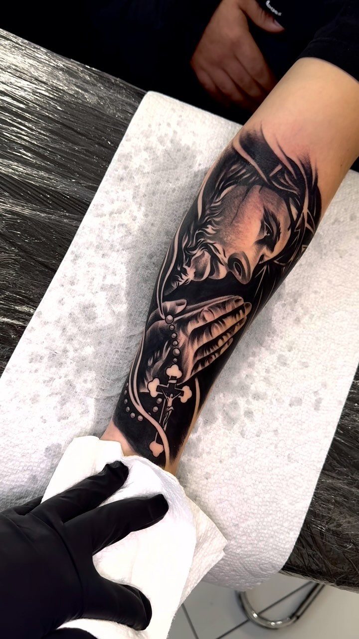 Black and grey forearm tattoo of praying Jesus with a rosary and cross.