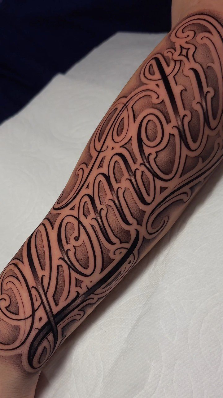 Ornate calligraphic lettering tattoo in black and grey ink on a person's forearm.