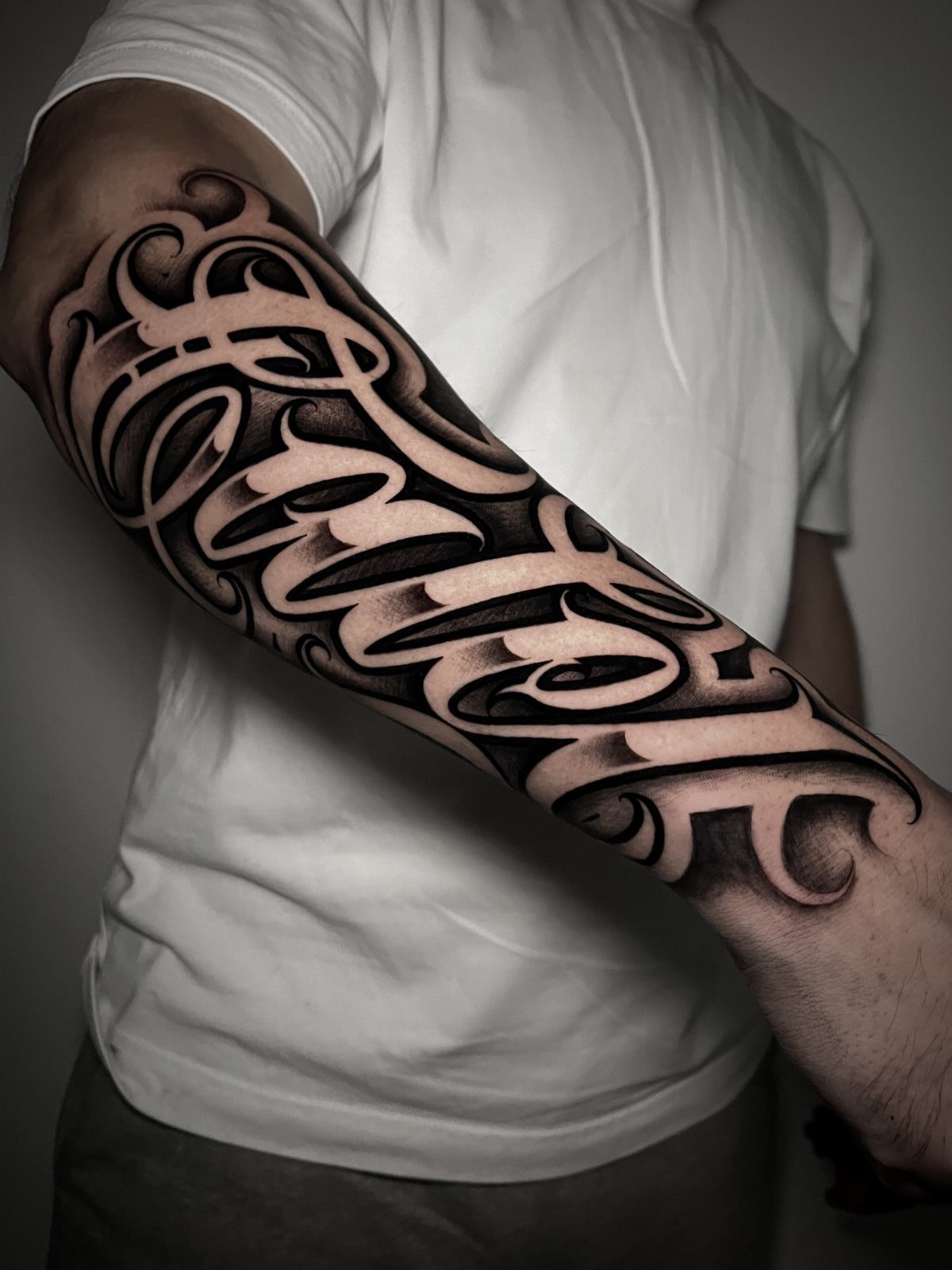 Stylized script tattoo on a forearm with flowing black and grey lettering.