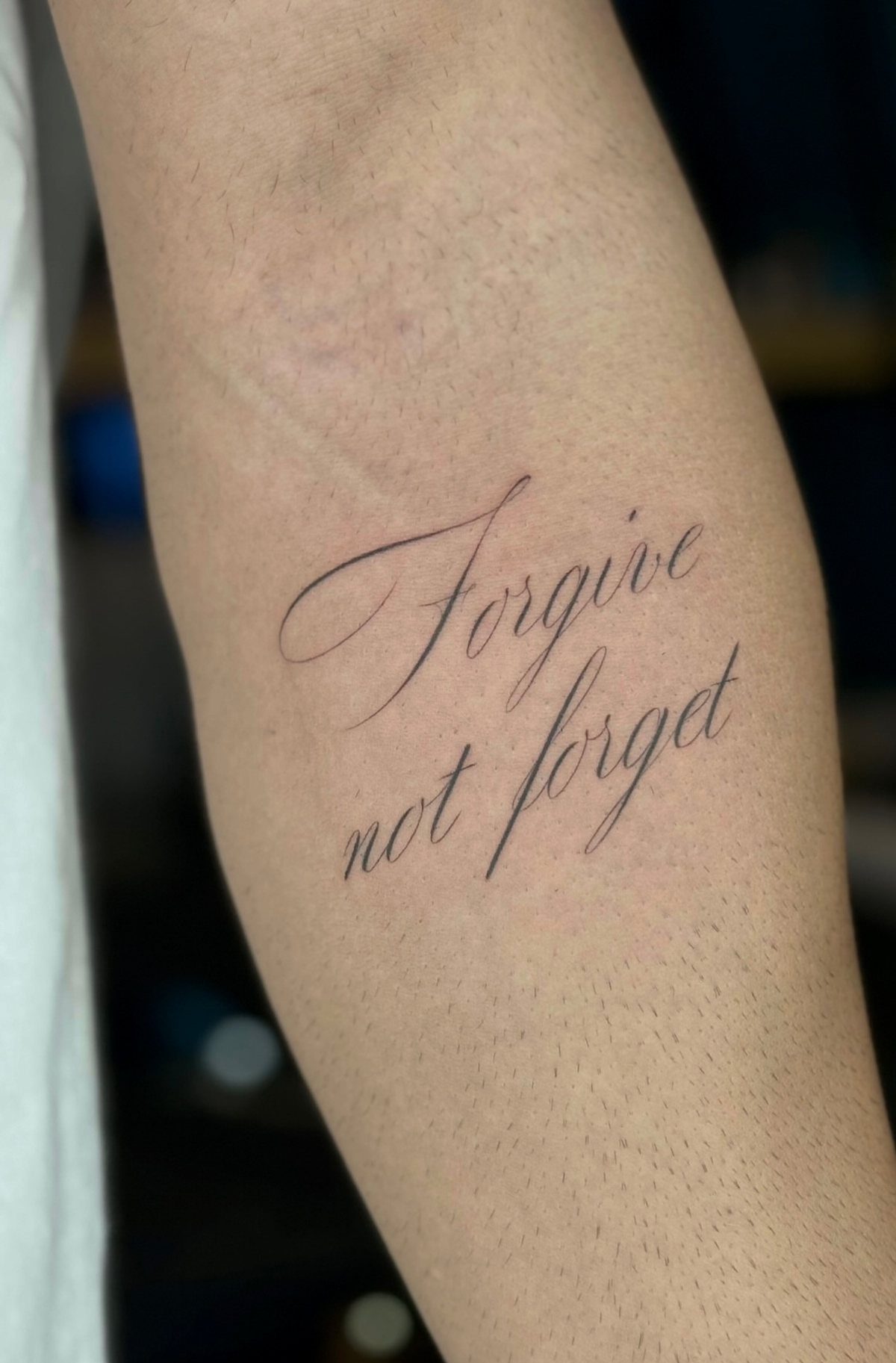 Cursive tattoo on a forearm reading Forgive not forget in fine line lettering.