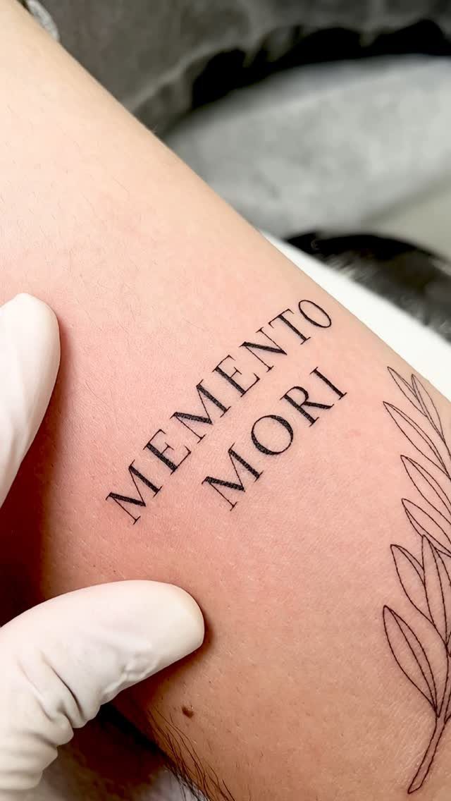 Serif font tattoo reading MEMENTO MORI with a leaf branch on skin