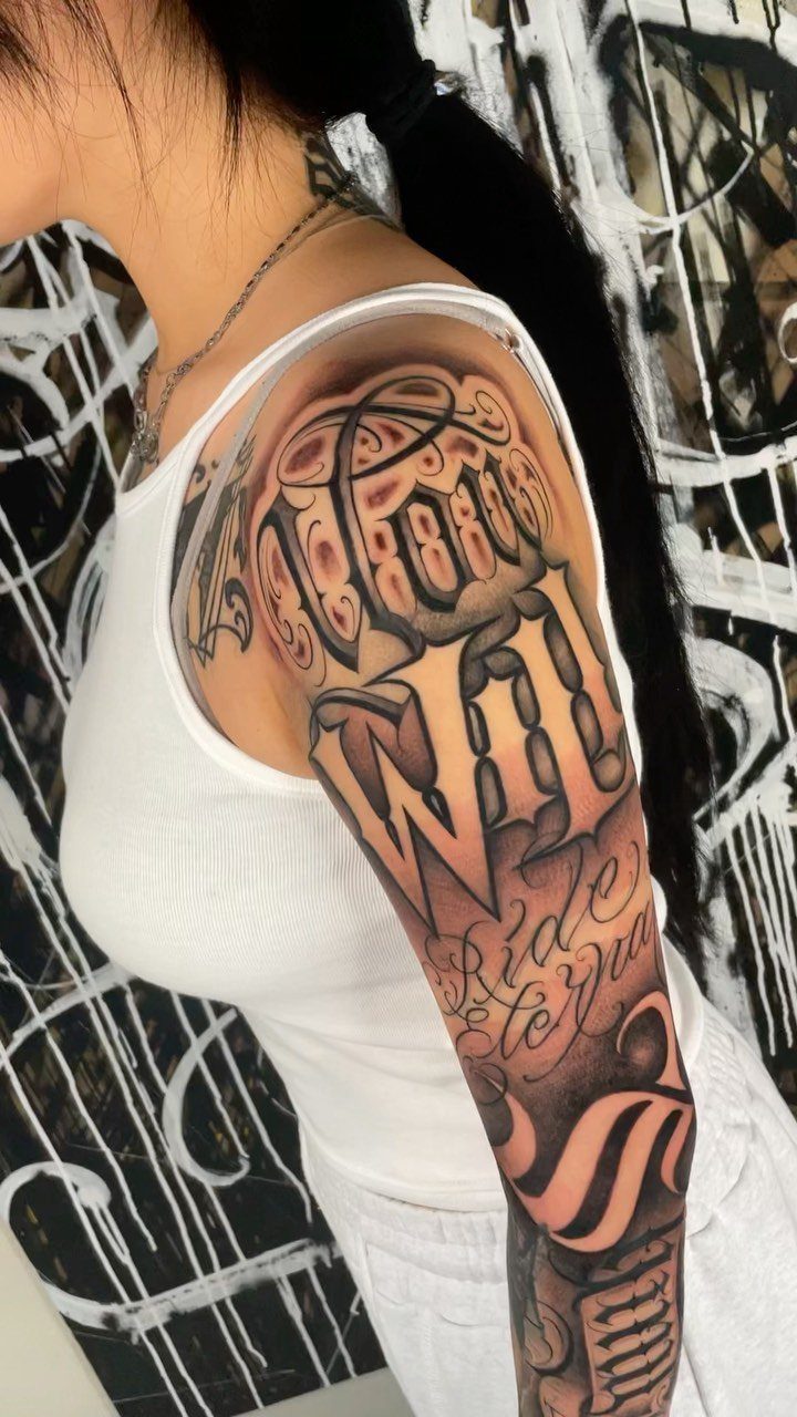 Ornate black and grey tattoo lettering on a person's upper arm and shoulder.
