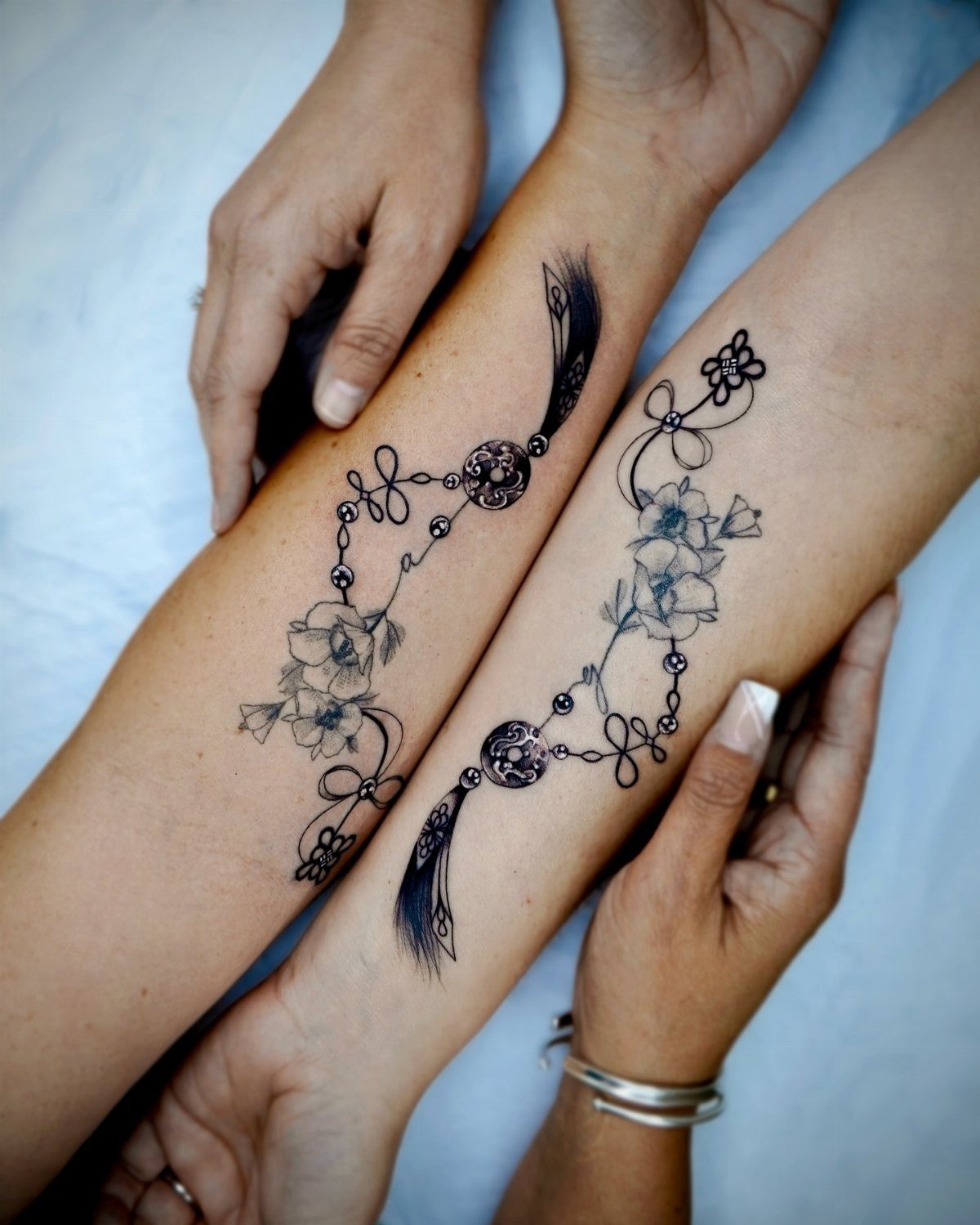 Matching black ink floral and tassel tattoos on two adjacent forearms.