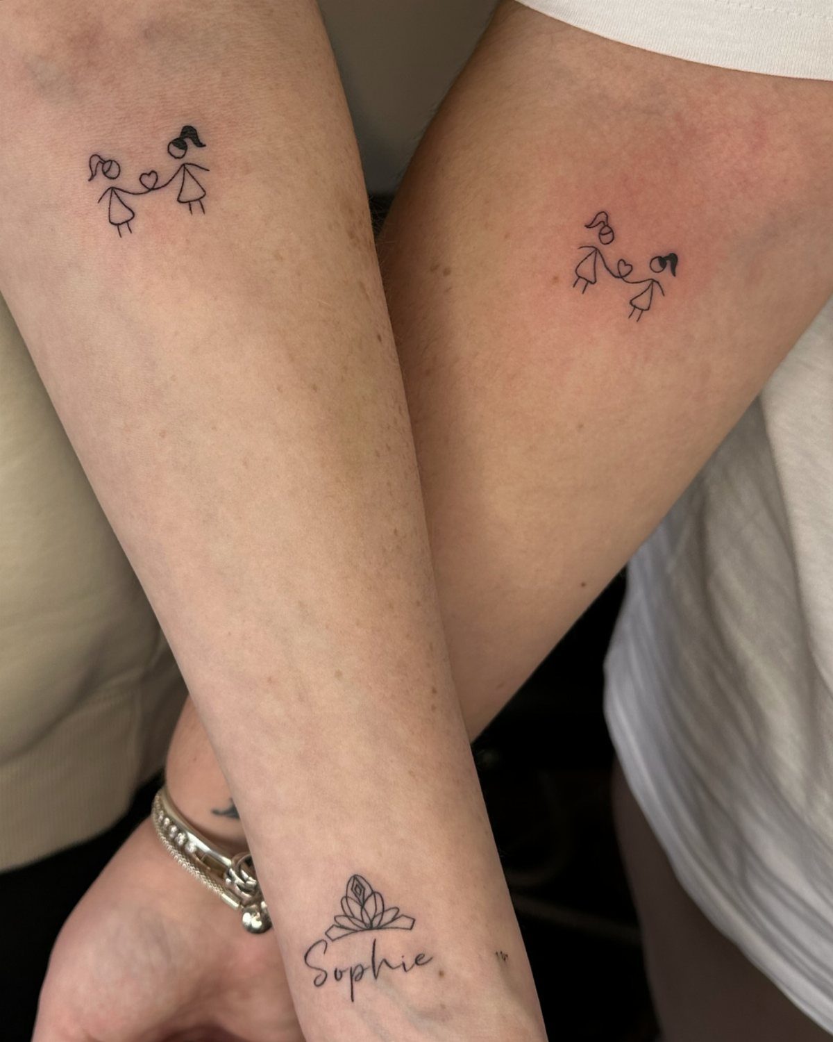 Two forearms with matching stick-figure girl tattoos holding hands in black ink.