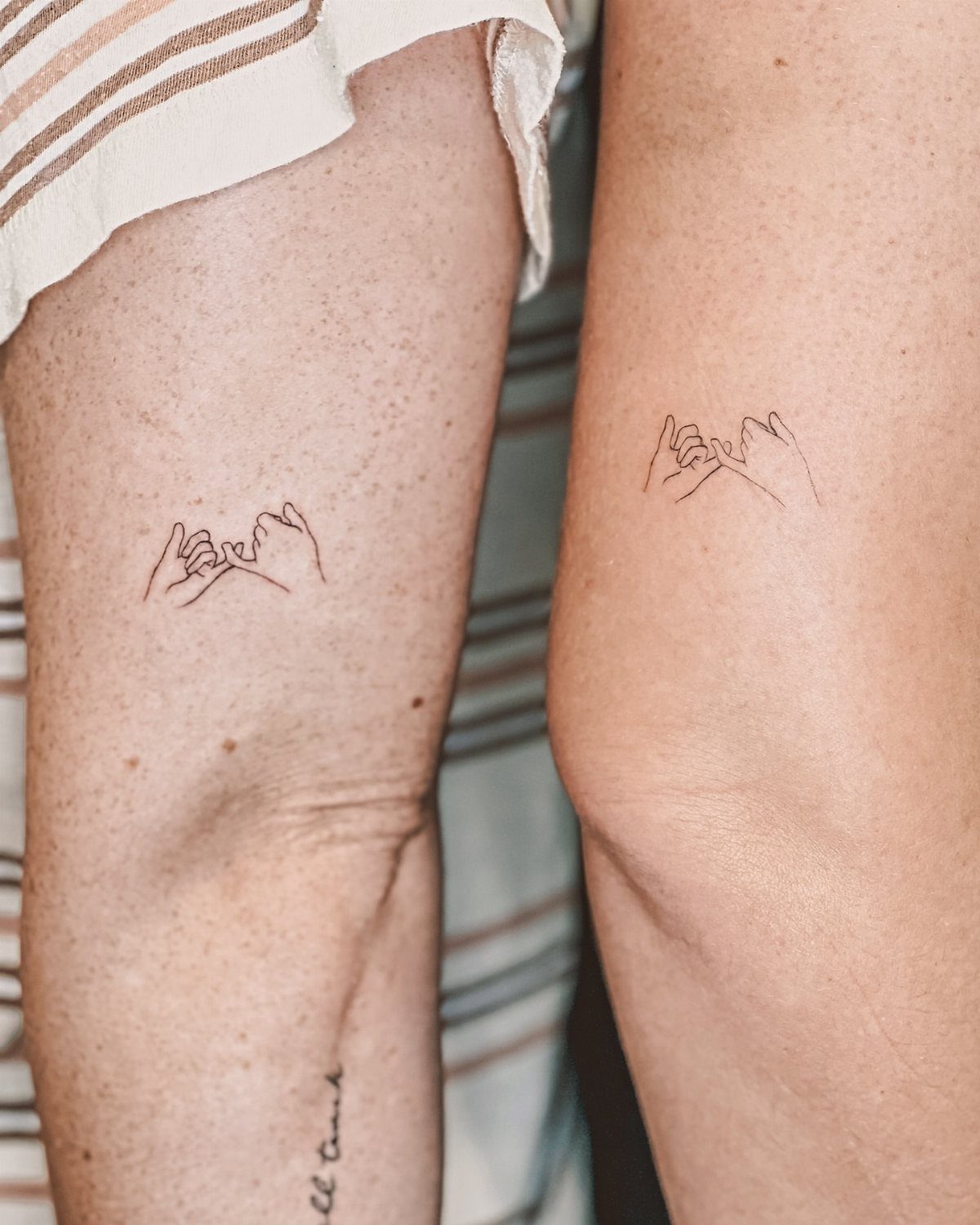 Matching pinkie promise tattoos on the legs of two people side by side.