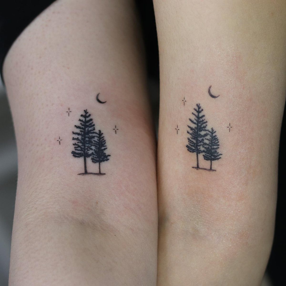 Two matching tattoos of pine trees and a moon on inner forearms.