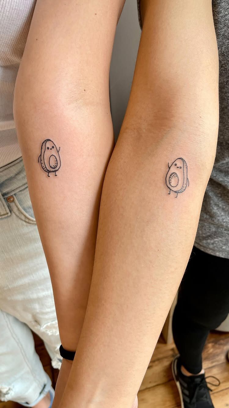 Two forearms with matching small avocado half tattoos featuring cute faces.