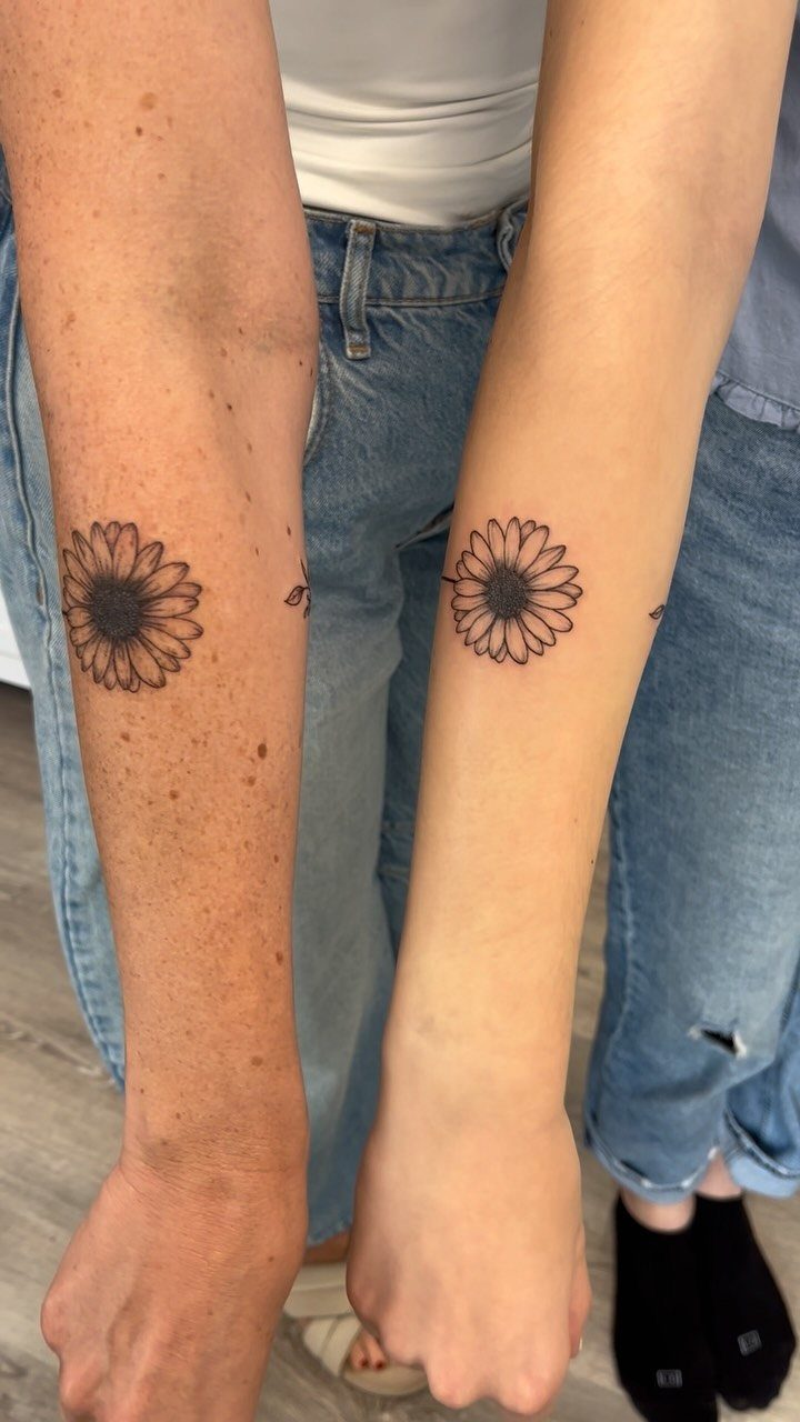 Matching black ink daisy tattoos on the inner forearms of two different people.