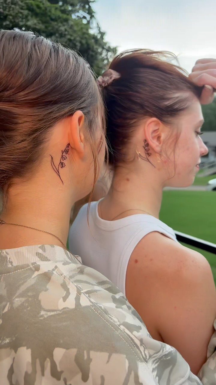 Two women showing matching small purple floral tattoos behind their ears outdoors.