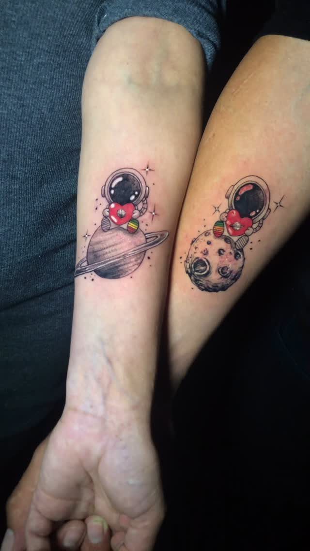 Matching tattoos of astronauts with hearts on a ringed planet and a moon.