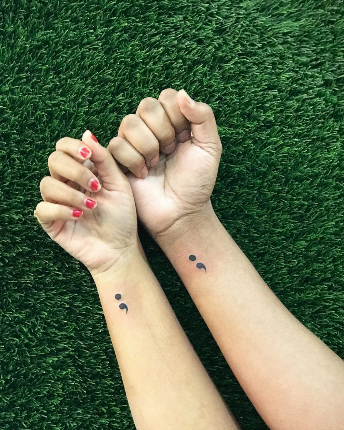 Two wrists with matching black semicolon tattoos resting on green artificial grass.