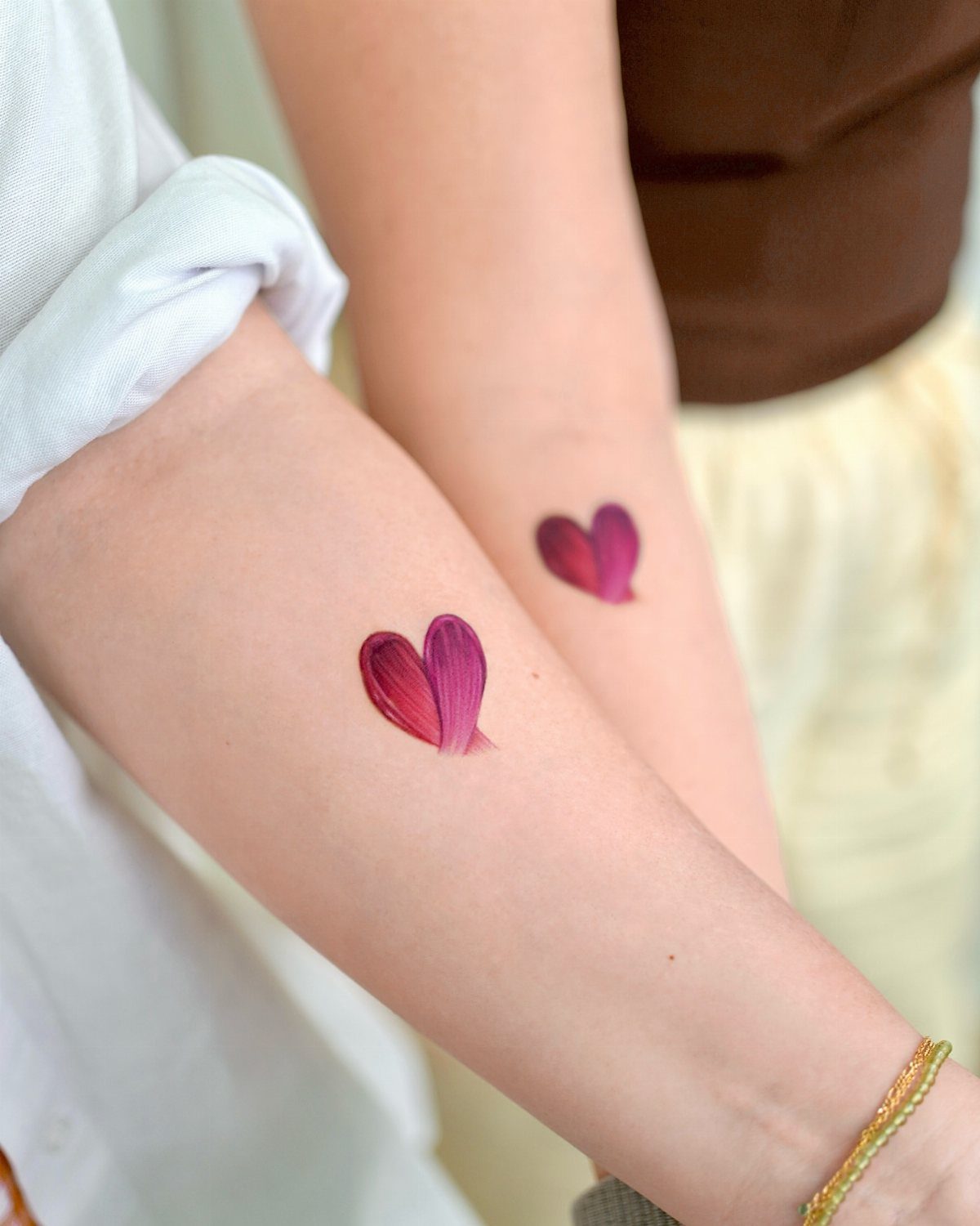 Two forearms with matching pink and purple watercolor heart tattoos on the wrists.