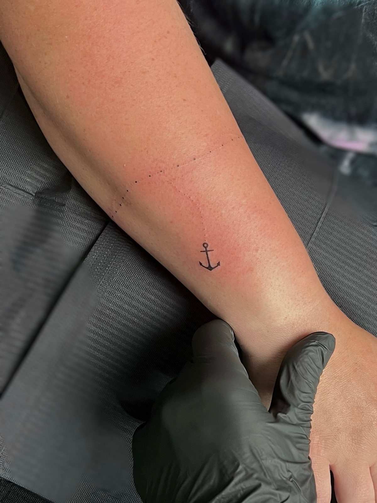 Small black anchor tattoo on a person's inner forearm held by gloved hand.