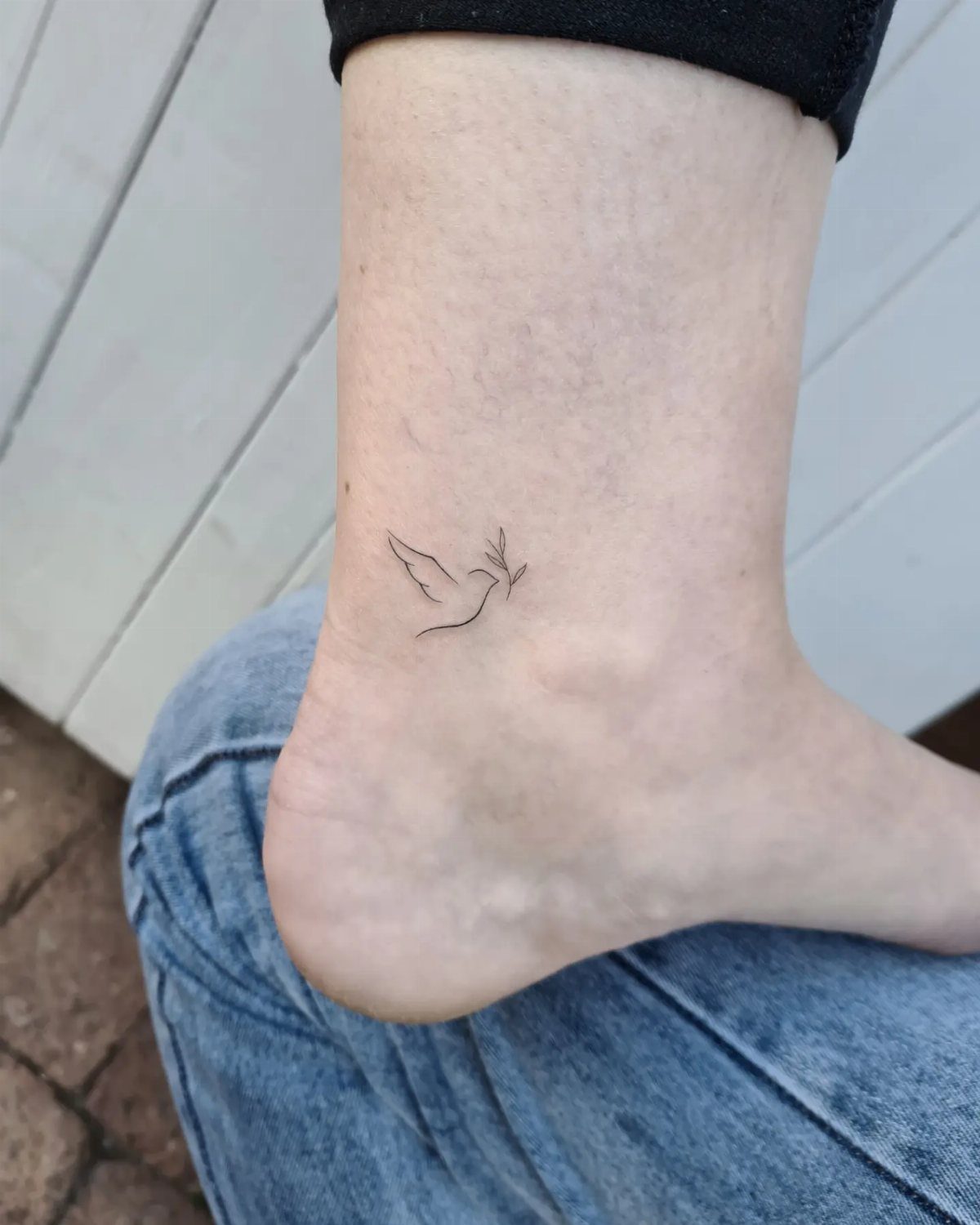 Small black ink bird tattoo on a person's ankle against white wood.
