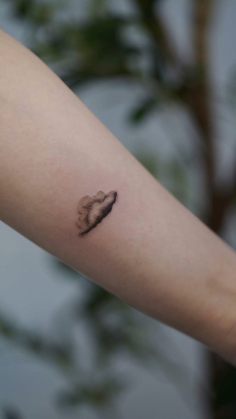 Small black ink cloud tattoo on a person's forearm with blurred greenery background