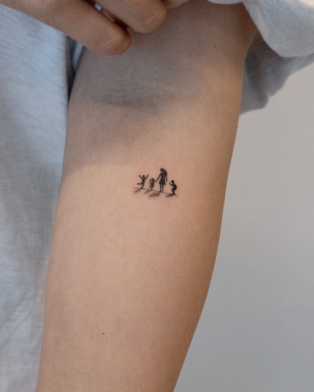 Small black silhouette tattoo of a parent and two children on a forearm.