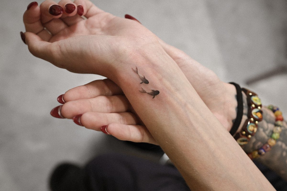 Two tiny black fish tattoos on a person's inner wrist with red nails.